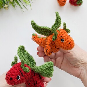 May include: Two crocheted stuffed toys shaped like a carrot and a strawberry. The carrot is orange with green leaves and the strawberry is red with green leaves. Both toys have black eyes and a smiling expression.