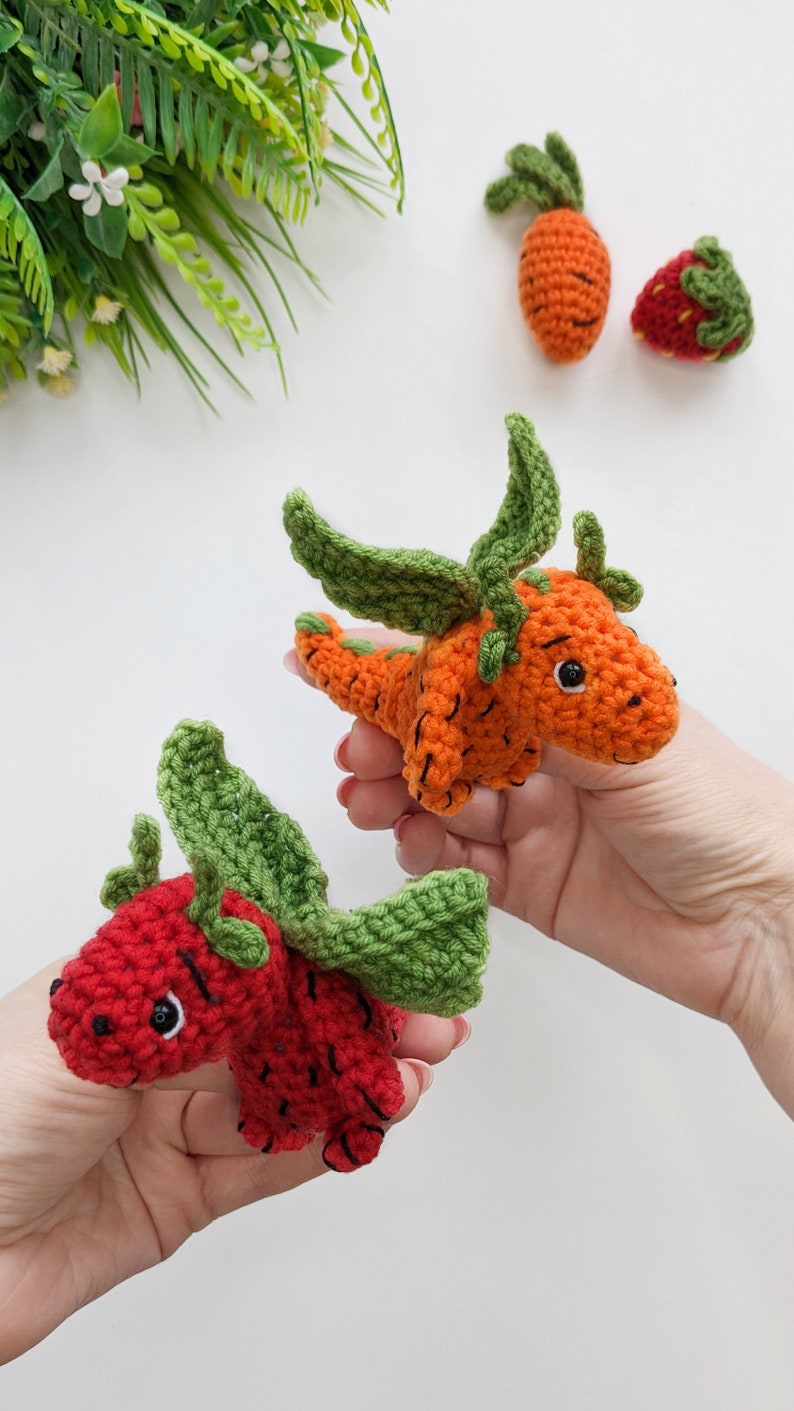 May include: Two crocheted stuffed toys shaped like a carrot and a strawberry. The carrot is orange with green leaves and the strawberry is red with green leaves. Both toys have black eyes and a smiling expression.