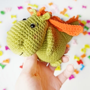 May include: A green crocheted dragon with orange wings. The dragon has black eyes and a small smile. The dragon is being held in a hand.