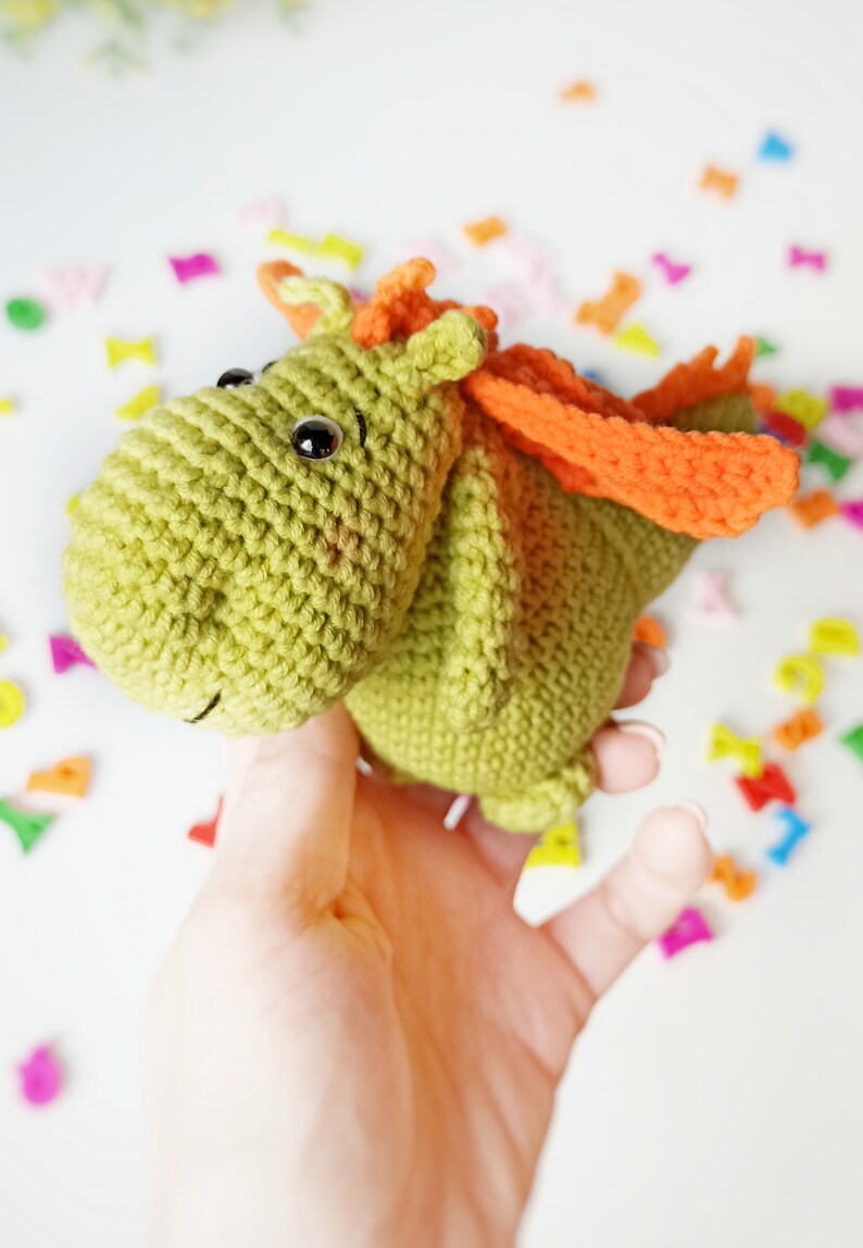 May include: A green crocheted dragon with orange wings. The dragon has black eyes and a small smile. The dragon is being held in a hand.