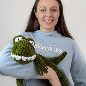 Puede incluir: Un peluche de cocodrilo verde tejido a ganchillo con dientes y ojos blancos. El peluche lo sostiene una persona que lleva una sudadera con capucha azul claro con el texto "Brooklyn USA". El cocodrilo tiene un aspecto suave y texturizado.