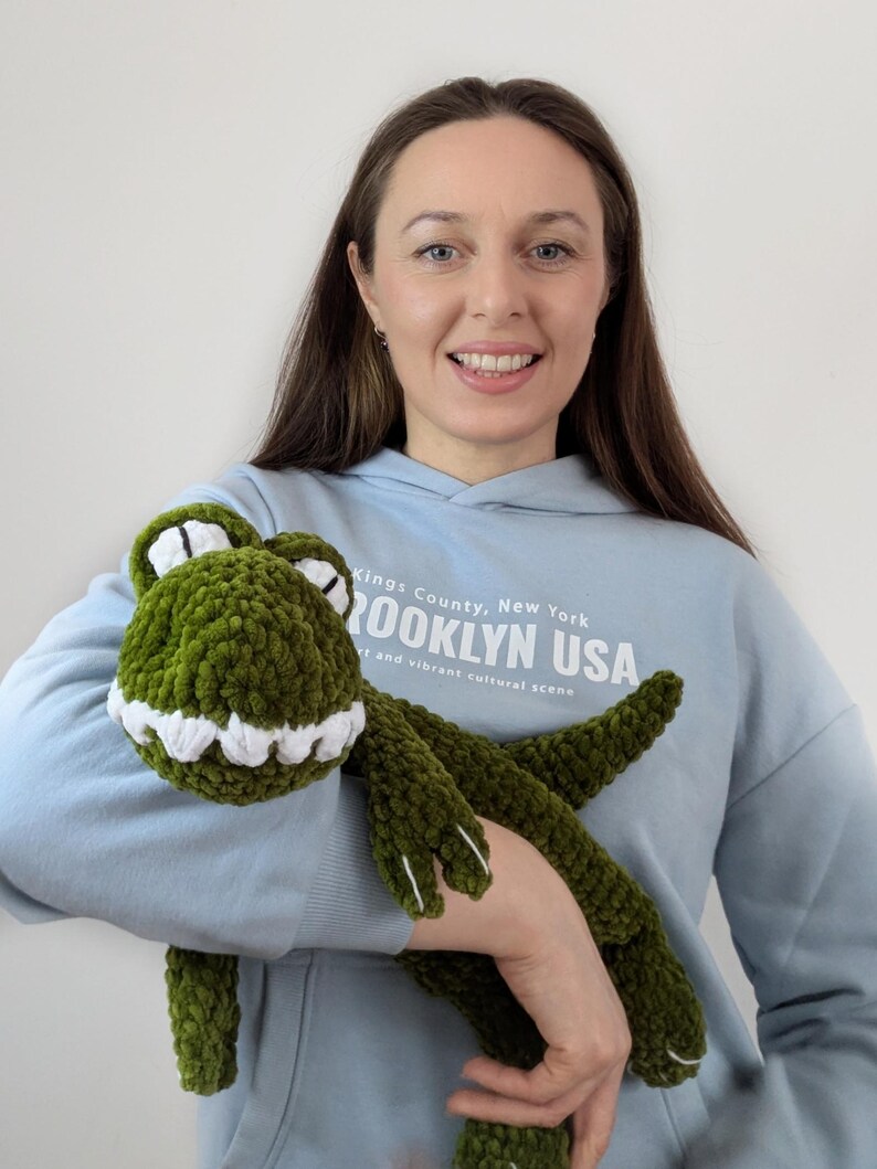 Puede incluir: Un peluche de cocodrilo verde tejido a ganchillo con dientes y ojos blancos. El peluche lo sostiene una persona que lleva una sudadera con capucha azul claro con el texto "Brooklyn USA". El cocodrilo tiene un aspecto suave y texturizado.