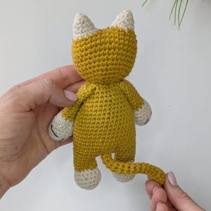K&ouml;nnte beinhalten: Eine handgefertigte H&auml;kelkatzenpuppe in Senfgelb mit cremefarbenen Akzenten. Die Katze hat einen runden K&ouml;rper, kleine Arme und Beine sowie einen langen Schwanz. Die Ohren und Pfoten sind cremefarben. Die Puppe wird von einer Person gehalten.