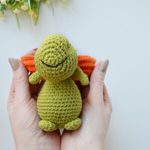 May include: A green crocheted turtle toy with orange crocheted wings. The turtle has a smiling face with black eyes and a black mouth.