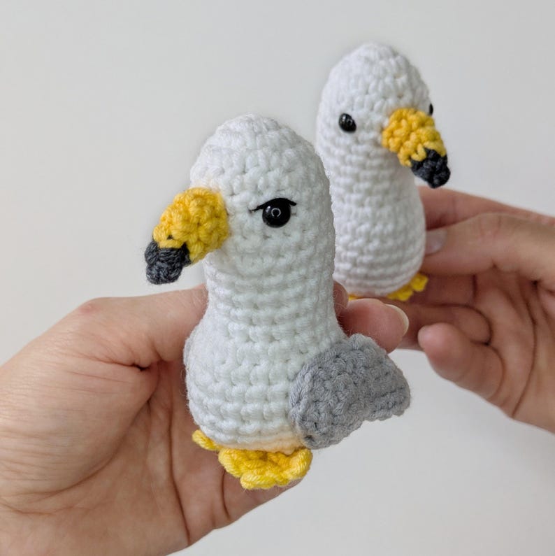 Op de afbeelding: Twee kleine, handgemaakte gehaakte meeuwen met lichamen van wit garen, grijze vleugels en felgele snavels met zwarte punten. Elke vogel heeft &eacute;&eacute;n zwart kraaloog aan de zijkant van de kop en kleine gele voetjes aan de onderkant.