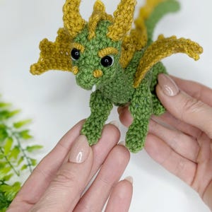May include: A small green crocheted dragon with yellow wings and black eyes. The dragon is being held in two hands.