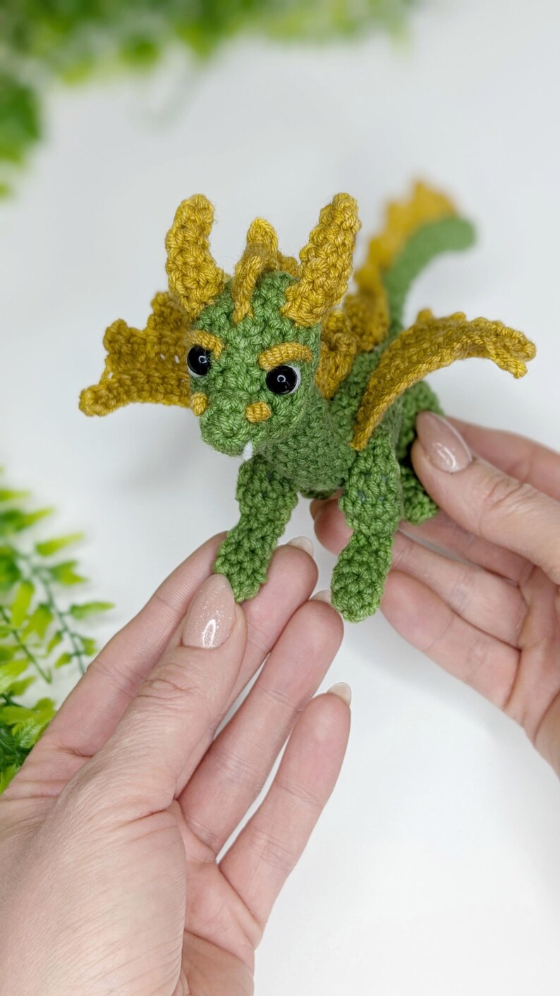 May include: A small green crocheted dragon with yellow wings and black eyes. The dragon is being held in two hands.