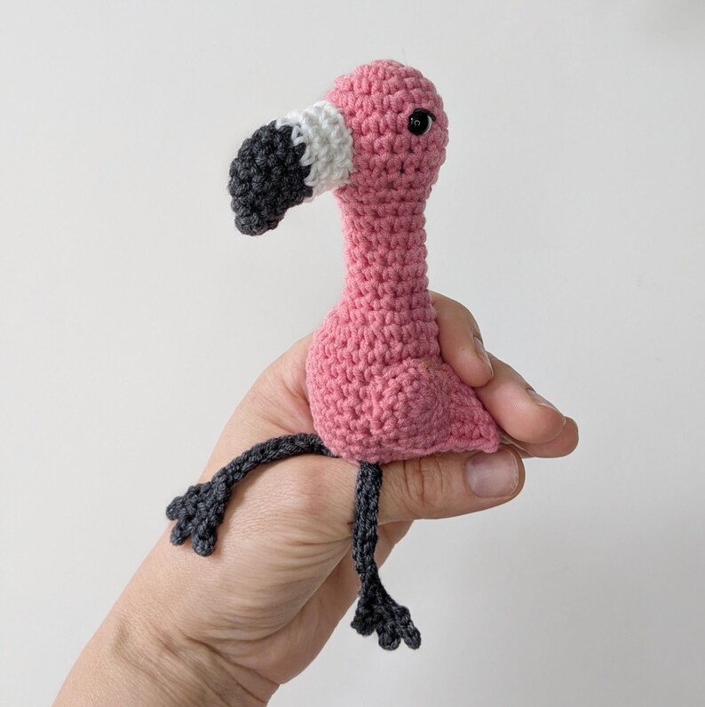 May include: A small, handmade crochet flamingo toy featuring a bright pink body, a long neck, and a curved beak with white and dark gray sections. The flamingo has two thin, dark gray crocheted legs with webbed feet and a single black eye.