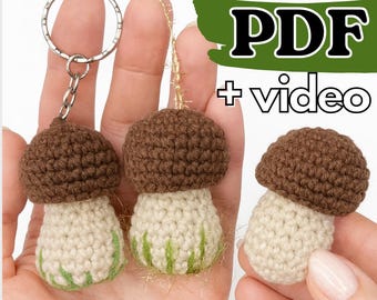 Può includere: Portachiavi e ornamenti a forma di fungo all'uncinetto. I funghi hanno cappelli marroni e gambi bianco sporco con accenti verdi. Un portachiavi ha un anello argentato. L'immagine include il testo "PDF + video".