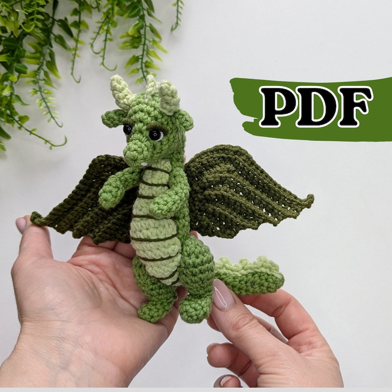 May include: Green crocheted dragon amigurumi toy with wings. The dragon has a light green body and dark green wings. The dragon has black eyes and a light green snout.