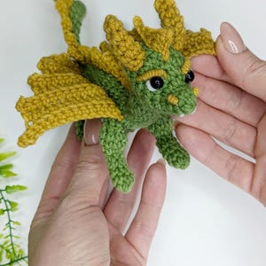 May include: A green crocheted dragon with yellow wings and black eyes. The dragon is small and is being held in the palms of two hands.