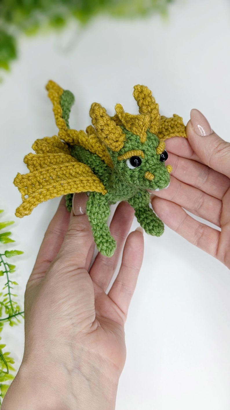 May include: A green crocheted dragon with yellow wings and black eyes. The dragon is small and is being held in the palms of two hands.