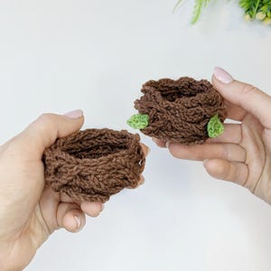 May include: Two handmade, brown crochet nests, each with a small green leaf accent. The nests are round and have a textured appearance, suggesting a cosy, natural theme. The image is well-lit, highlighting the details of the yarn and the craftsmanship.