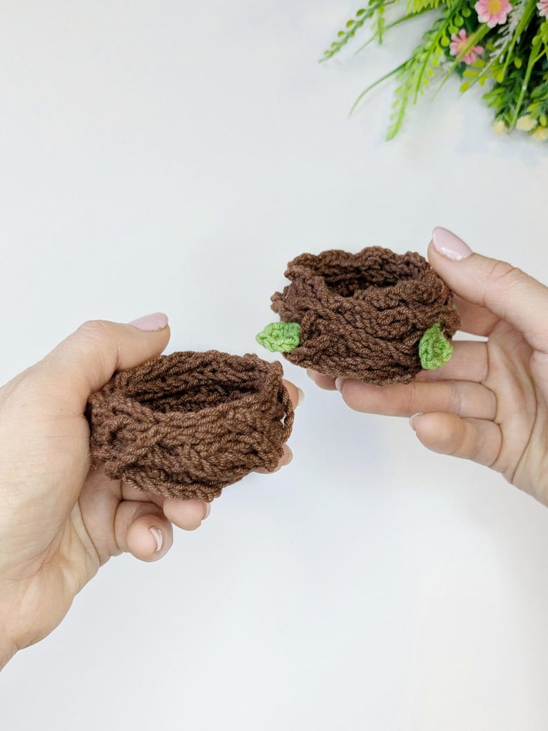 May include: Two handmade, brown crochet nests, each with a small green leaf accent. The nests are round and have a textured appearance, suggesting a cosy, natural theme. The image is well-lit, highlighting the details of the yarn and the craftsmanship.