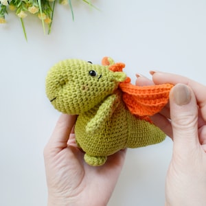 May include: A green crocheted dragon toy with orange wings. The dragon has black eyes and a small smile.