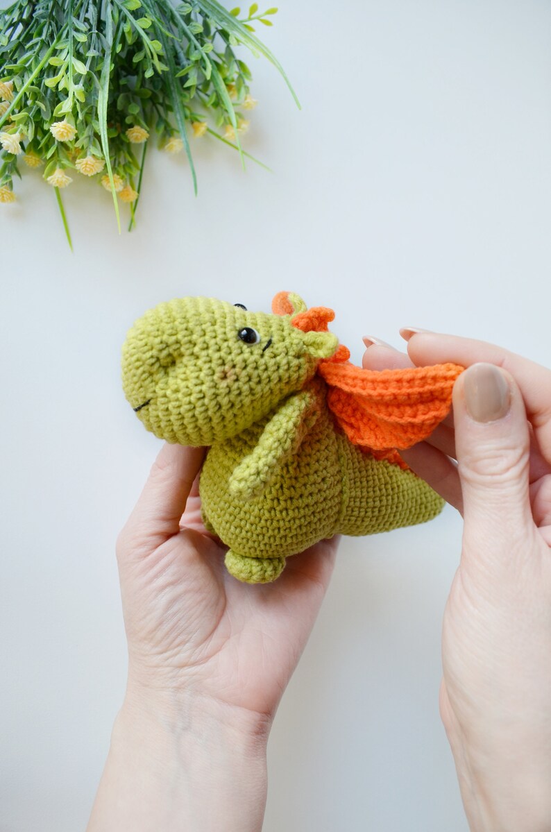 May include: A green crocheted dragon toy with orange wings. The dragon has black eyes and a small smile.