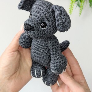 K&ouml;nnte beinhalten: Ein kleines, dunkelgraues H&auml;kelhund-Spielzeug mit strukturierter Garnoberfl&auml;che, Schlappohren, einer schwarz gestickten Nase und runden schwarzen Kunststoffaugen. Die Pl&uuml;schfigur hat einen kurzen Schwanz und kleine Pfoten mit wei&szlig;en Details.