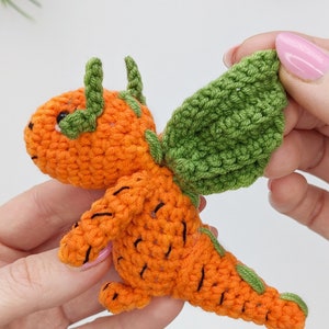 May include: A crocheted carrot-shaped dragon with green wings. The dragon is orange with black details and has a small, round head with black eyes.