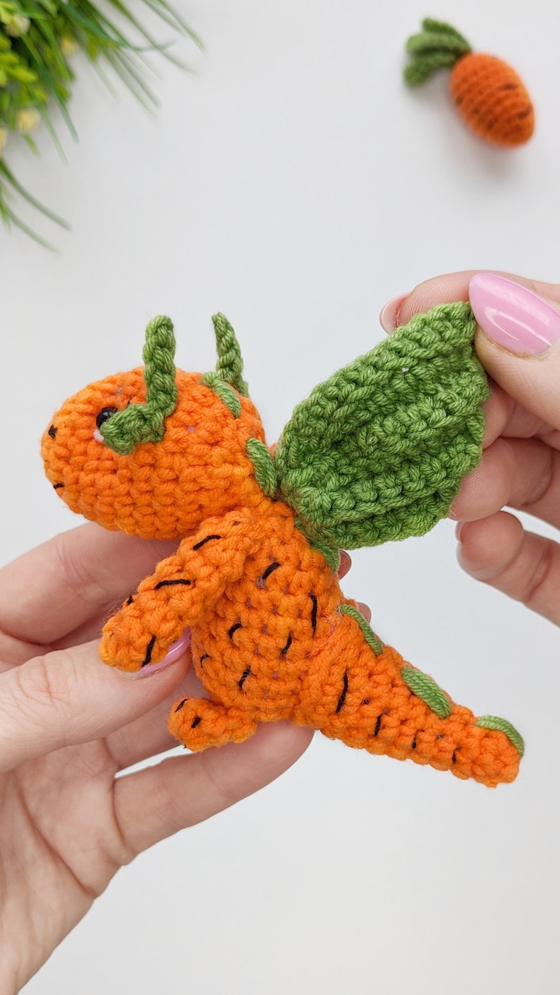 May include: A crocheted carrot-shaped dragon with green wings. The dragon is orange with black details and has a small, round head with black eyes.