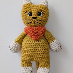 May include: A mustard yellow crocheted cat doll with cream-colored ears and paws. The cat features black plastic safety eyes, embroidered black whiskers, and a small black nose. It wears a textured orange triangular bandana around its neck.