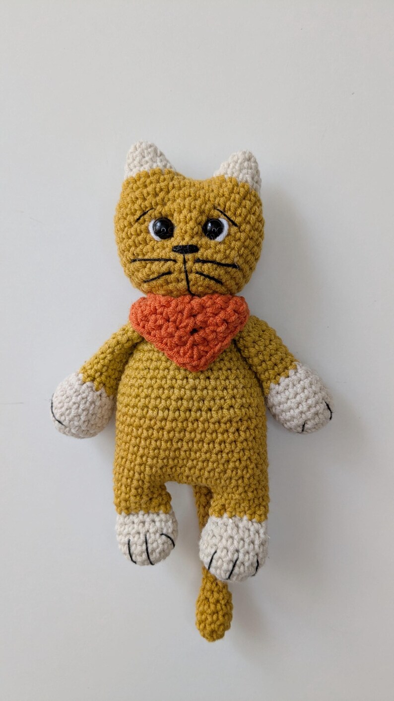 May include: A mustard yellow crocheted cat doll with cream-colored ears and paws. The cat features black plastic safety eyes, embroidered black whiskers, and a small black nose. It wears a textured orange triangular bandana around its neck.