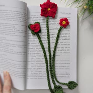 Puede incluir: Un marcap&aacute;ginas de ganchillo con tres tallos verdes largos y peque&ntilde;as hojas verdes en la parte inferior. Cada tallo est&aacute; rematado con una flor roja con el centro amarillo. El marcap&aacute;ginas est&aacute; colocado dentro de un libro abierto.