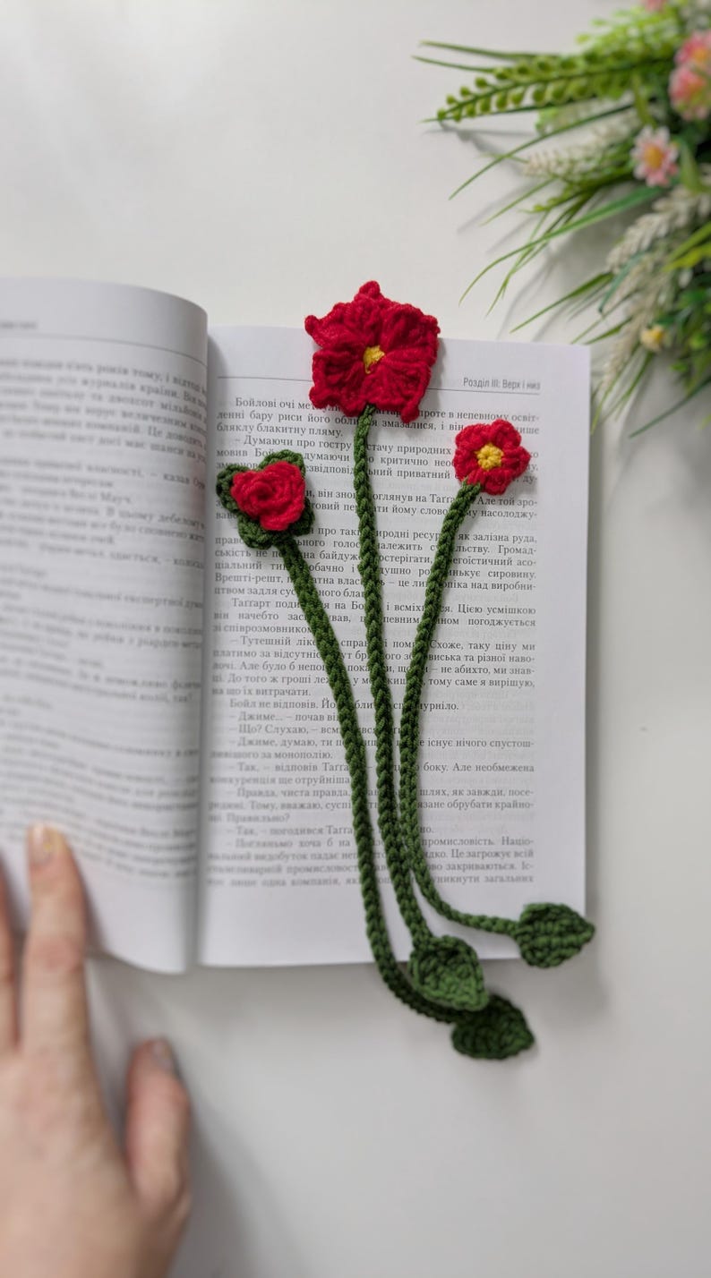 Puede incluir: Un marcap&aacute;ginas de ganchillo con tres tallos verdes largos y peque&ntilde;as hojas verdes en la parte inferior. Cada tallo est&aacute; rematado con una flor roja con el centro amarillo. El marcap&aacute;ginas est&aacute; colocado dentro de un libro abierto.