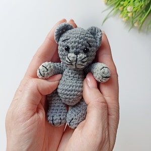 May include: A small gray crocheted cat toy with black eyes and a black nose. The cat is being held in a person's hands.