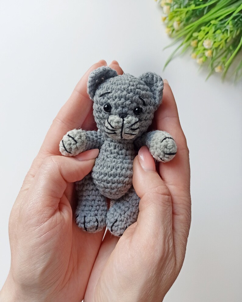 May include: A small gray crocheted cat toy with black eyes and a black nose. The cat is being held in a person's hands.
