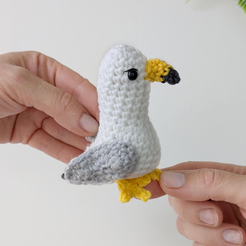 Op de afbeelding: Een klein, handgemaakt gehaakt meeuwspeeltje met een witte kop en lichaam, lichtgrijze vleugels, een gele snavel met een zwarte punt en gele pootjes. De vogel heeft &eacute;&eacute;n zwart kraaloog aan de zijkant van de kop, gemaakt met textuursteken.