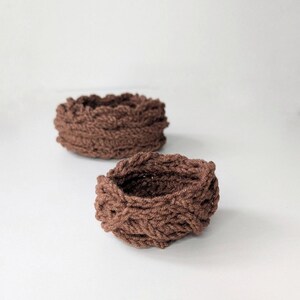 May include: Two small, round, brown woven baskets are shown against a white backdrop. The baskets have a textured, braided appearance, with a slightly uneven rim. The colour is a warm, earthy brown, suggesting a natural fibre material.