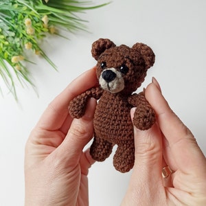 May include: A small brown crocheted teddy bear with black eyes and a brown nose. The bear is being held by two hands.