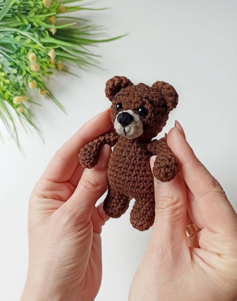 May include: A small brown crocheted teddy bear with black eyes and a brown nose. The bear is being held by two hands.
