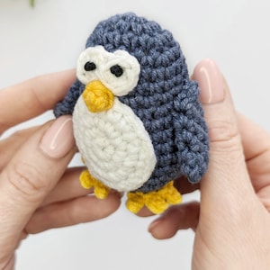 Pode incluir: Um brinquedo de pinguim de croch&ecirc; com corpo azul, barriga branca, bico e p&eacute;s amarelos e olhos pretos. O pinguim est&aacute; sendo segurado por duas m&atilde;os.