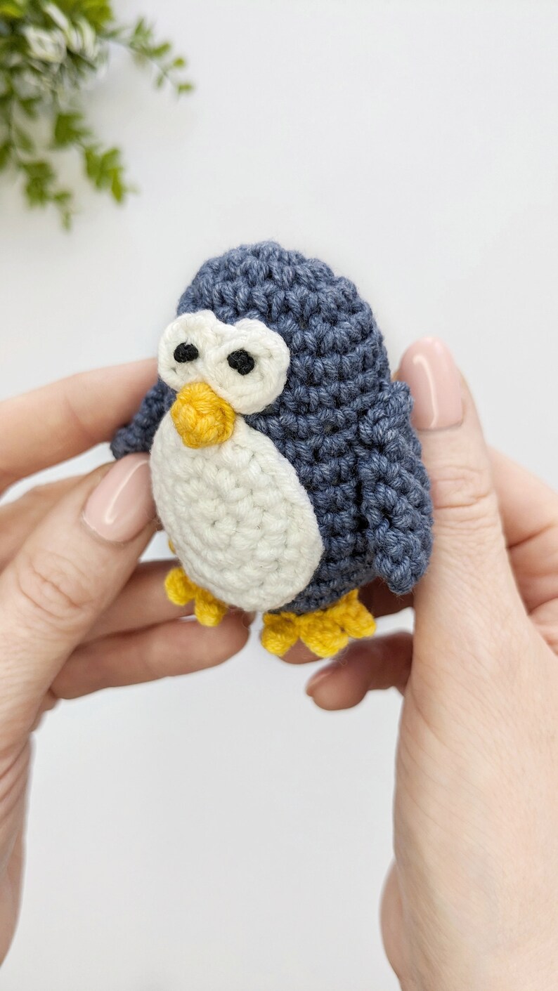 Pode incluir: Um brinquedo de pinguim de croch&ecirc; com corpo azul, barriga branca, bico e p&eacute;s amarelos e olhos pretos. O pinguim est&aacute; sendo segurado por duas m&atilde;os.