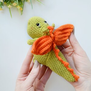 May include: A green crocheted dragon with orange wings. The dragon has a cute face with black eyes and a small smile.