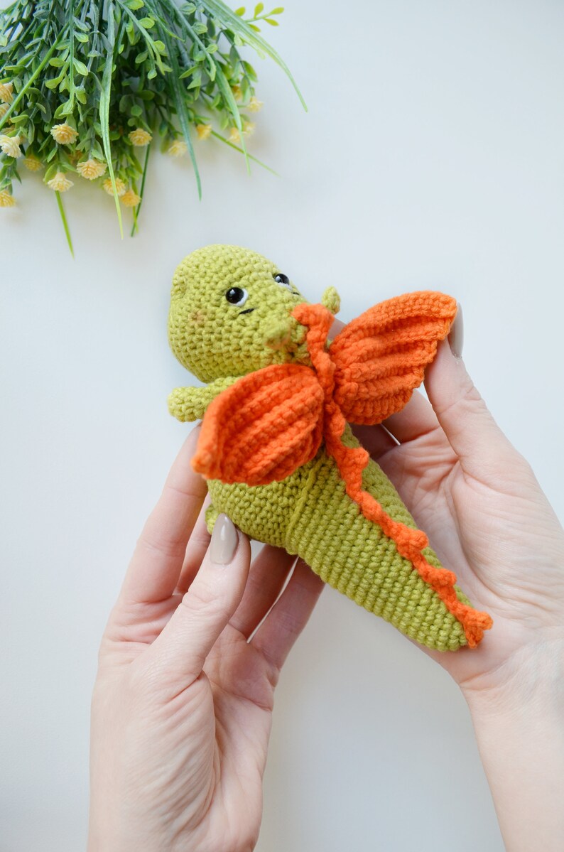 May include: A green crocheted dragon with orange wings. The dragon has a cute face with black eyes and a small smile.