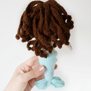 May include: A crocheted mermaid doll with brown hair and a light blue tail. The doll is being held by a hand.