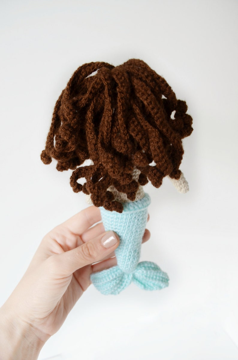 May include: A crocheted mermaid doll with brown hair and a light blue tail. The doll is being held by a hand.