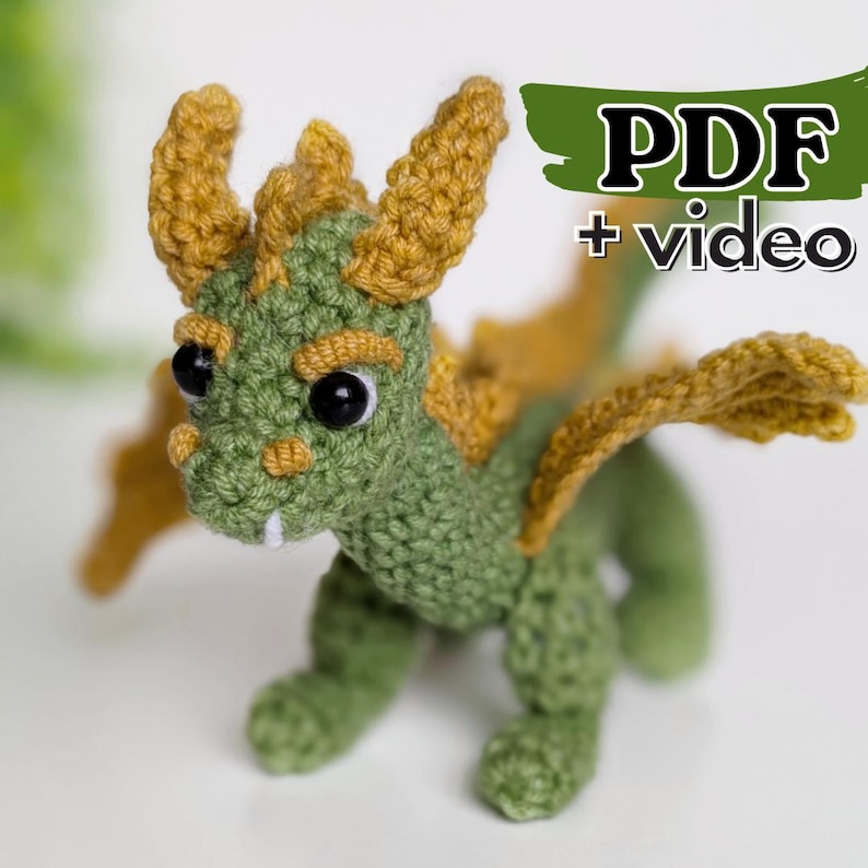 May include: A green and yellow crocheted dragon amigurumi toy with large black eyes. The toy has wings and a small mouth with white teeth. The image includes the text "PDF + video".