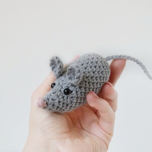 May include: A gray crocheted mouse toy with black eyes and a pink nose. The mouse is being held in a hand.