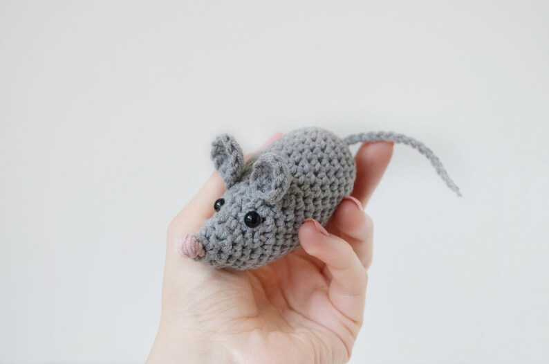 May include: A gray crocheted mouse toy with black eyes and a pink nose. The mouse is being held in a hand.