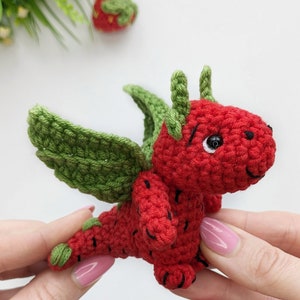 May include: A small crocheted strawberry dragon with green leaves for wings and horns. The dragon is red with black stitching for details and has a black eye.