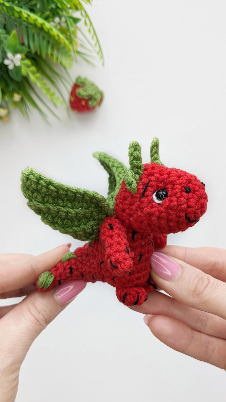 May include: A small crocheted strawberry dragon with green leaves for wings and horns. The dragon is red with black stitching for details and has a black eye.