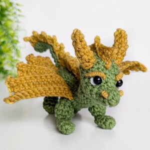 May include: A small green crocheted dragon with yellow wings and black eyes. The dragon is standing on a white surface.
