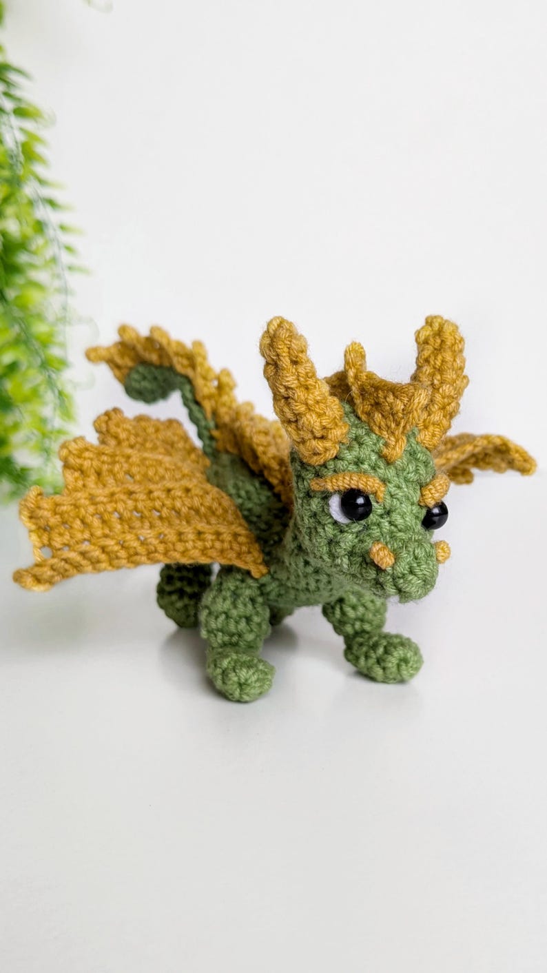 May include: A small green crocheted dragon with yellow wings and black eyes. The dragon is standing on a white surface.