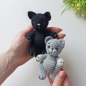 May include: Two crocheted cat toys, one black and one gray, are held in the hands of a person. The black cat has white stitching around its eyes and nose. The gray cat has white stitching around its eyes and nose and white paws.