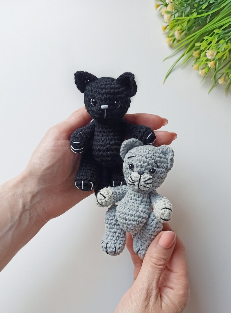 May include: Two crocheted cat toys, one black and one gray, are held in the hands of a person. The black cat has white stitching around its eyes and nose. The gray cat has white stitching around its eyes and nose and white paws.