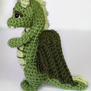 May include: A green crocheted dragon with a dark green crocheted cape. The dragon has a light green head and a light green tail.