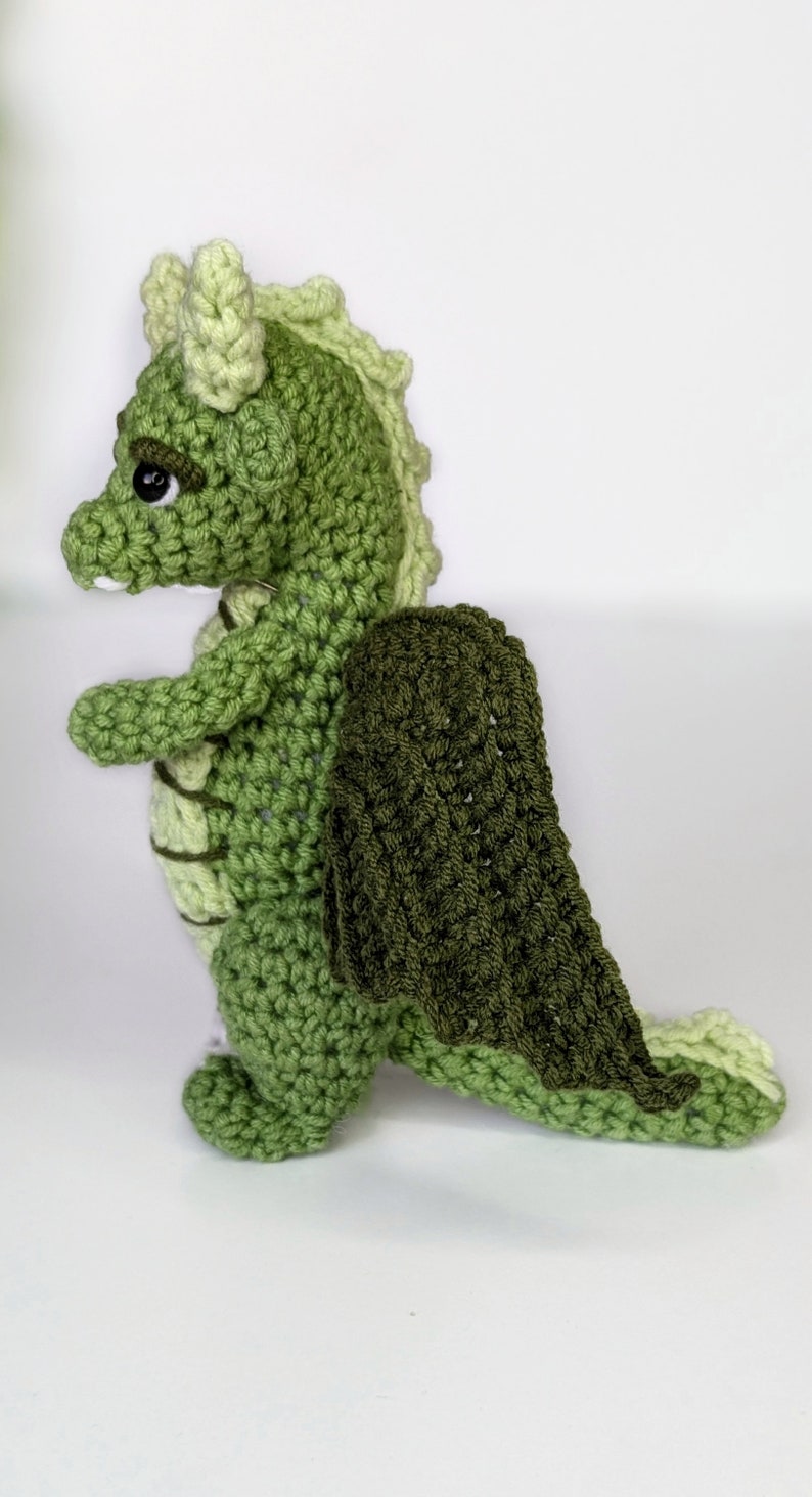 May include: A green crocheted dragon with a dark green crocheted cape. The dragon has a light green head and a light green tail.